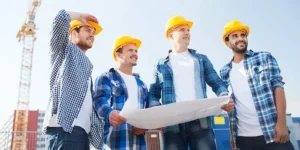 Four general contractors from The Mudacking Contractor in hard hats are standing together looking at a job site with a large crane in the background